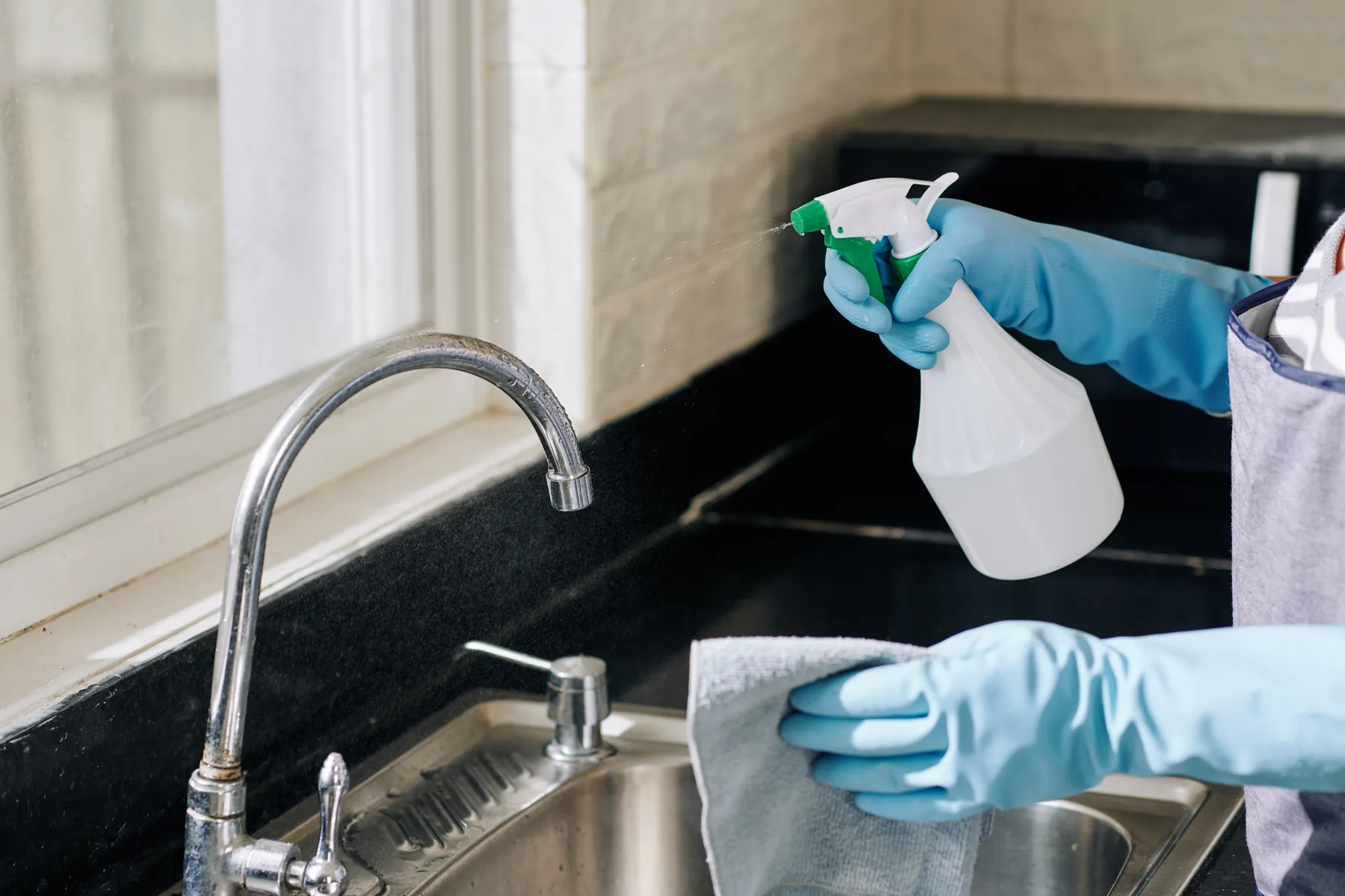 A cleaner in rubber gloves spraying and wiping down a kitchen worktop beside the sink